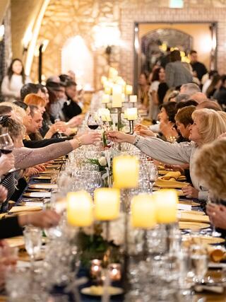 A long table filled with people celebrating, raising glasses in toasts, surrounded by candles and an elegant ambiance.