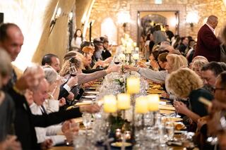 A large gathering of people toasting at a long dinner table, adorned with candles and elegant table settings, exuding a warm, celebratory atmosphere.