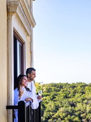 A couple enjoys a moment on a balcony, sipping drinks and smiling, with a lush green landscape and clear blue skies in the background.