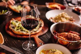 A table filled with a variety of dishes, including pizza, pasta, and multiple sides, accompanied by a glass of red wine.