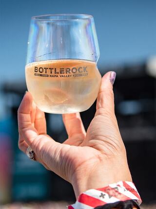 A hand holds a clear glass filled with white wine, featuring a label that reads "Bottlerock Napa Valley," against a bright blue sky.