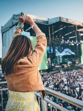 A woman with long hair raises her arms in excitement at a vibrant outdoor concert, surrounded by a large crowd and bright stage lights.