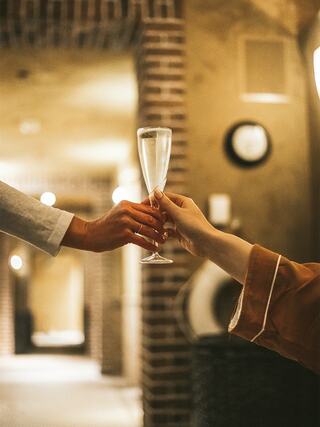 Two hands toast with champagne flutes in a cozy, softly lit corridor, suggesting a moment of celebration and connection.