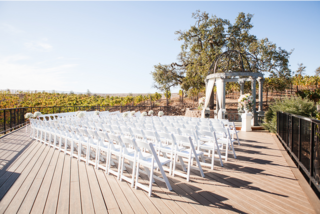 An elegant outdoor wedding venue with white chairs arranged neatly in front of a floral-adorned gazebo, surrounded by vineyards.