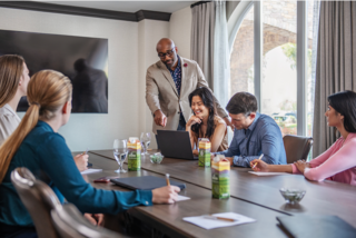 A diverse group of professionals engages in a meeting, with one speaker presenting while others listen and discuss, surrounded by refreshments.