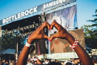 Hands form a heart shape in front of a vibrant concert stage at the BottleRock festival, capturing the excitement and energy of the crowd.