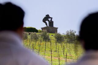 A vineyard scene features a statue of a figure working, with people in the foreground observing the lush landscape and grapevines.