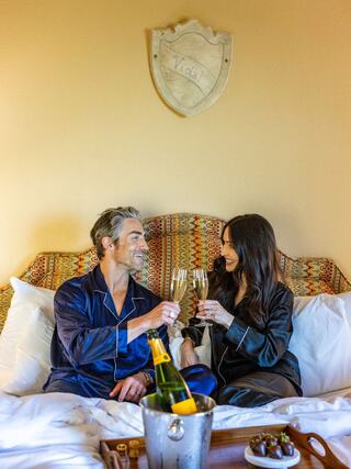 A couple in pajamas clinks champagne glasses while sitting on a bed adorned with a colorful headboard, sharing a cozy moment.