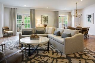 A cozy, modern living area features a spacious gray sofa, circular coffee table, elegant chandelier, and large windows with natural light.