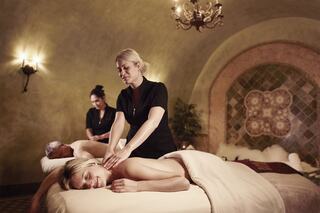 A serene spa setting with two clients receiving massages from skilled therapists, creating a relaxing atmosphere illuminated by soft lighting.