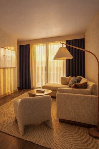 A cozy living space features a light-toned sofa, stylish armchair, and warm lighting from large windows, creating a welcoming atmosphere.