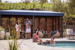 Four friends enjoy drinks by a pool with vineyard in the background, relaxing under a shaded area surrounded by lush greenery and cozy seating.