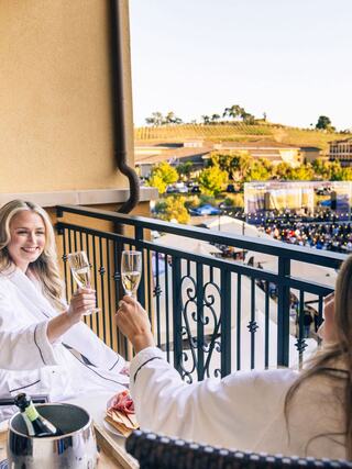 Two women in bathrobes toast with glasses of champagne on a balcony, overlooking a scenic vineyard and festive gathering below.