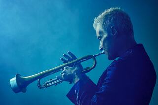 A musician plays a trumpet, silhouetted against a blue, smoky backdrop, exuding intensity and passion.