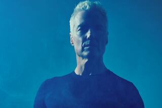 A silhouette of a man with light hair, facing forward against a smoky blue background, exuding a mysterious and striking presence.