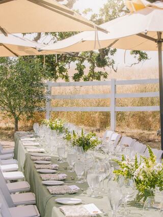 A beautifully set outdoor dining table adorned with glasses and floral arrangements, shaded by large umbrellas, surrounded by nature.