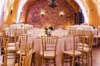 An elegant dining setup with gold chairs and ornate tables under warm lighting, featuring a stone wall and chandeliers. Perfect for events.