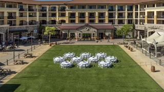 An outdoor venue features neatly arranged round tables on grass, surrounded by a charming hotel and patio seating under clear skies.