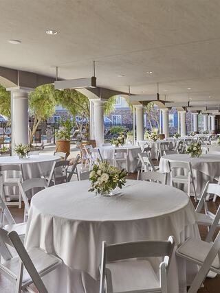 An elegant event space features round tables with white linens, adorned with floral centerpieces, surrounded by greenery and columns.
