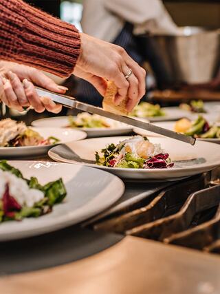 A hand slices bread over elegantly plated salads, showcasing a careful and artistic presentation in a bustling kitchen.