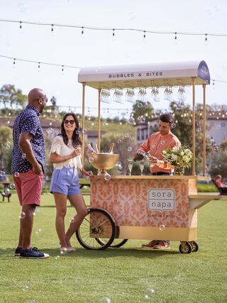 A vibrant outdoor scene features a bubble cart where a couple engages with a vendor, enjoying a lively atmosphere on a sunny day.