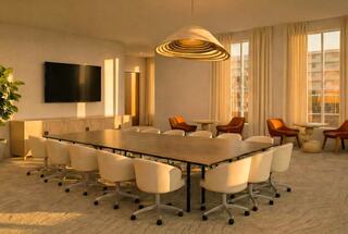 A modern conference room features a long table surrounded by white chairs, a large screen, cozy seating, and warm natural light.