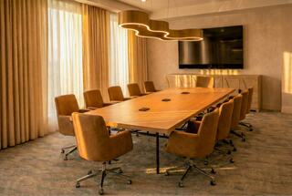 A modern conference room with a long wooden table, plush brown chairs, large windows, and stylish light fixtures.