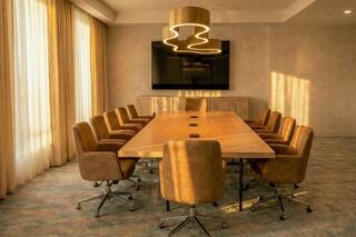 A modern conference room features a large wooden table surrounded by upholstered chairs, with soft natural light streaming through sheer curtains.