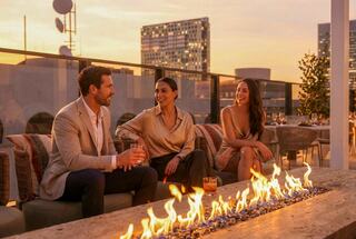 Three elegantly dressed friends enjoy drinks and conversation by a cozy outdoor fire pit, with a sunset skyline in the background.