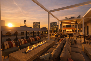 A stylish rooftop lounge features cozy seating around a glowing fire pit, with a stunning sunset and city skyline in the background.