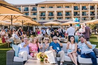 A group of five people smile and toast on a couch at an outdoor event, with a crowd and elegant setting in the background.