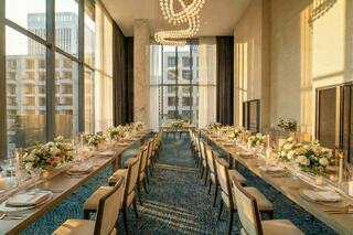 An elegantly set long dining table with floral arrangements, surrounded by modern decor and large windows letting in warm sunlight.