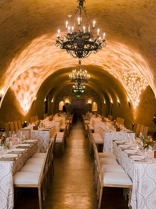 An elegant banquet hall features long tables adorned with tableware, surrounded by soft lighting and ornate chandeliers under a vaulted ceiling.