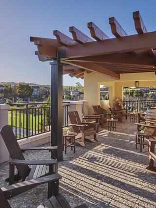 A serene patio features wooden chairs under a pergola, offering a peaceful view of greenery and a sunny sky. Perfect for relaxation.