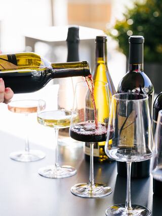 A hand pours red wine into a glass, surrounded by assorted wine bottles and various other glasses, creating a lively setting.
