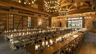 A rustic, warmly lit banquet hall features long tables adorned with candles and modern chandeliers, creating an inviting atmosphere.
