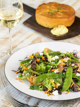 A vibrant salad with mixed greens, snap peas, carrots, and feta cheese is served alongside cornbread and a glass of white wine.