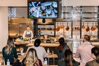 A culinary instructor demonstrates cooking techniques to an engaged audience in a well-equipped kitchen setting.