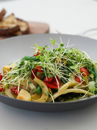 A vibrant pasta dish featuring colorful vegetables, fresh sprouts, and herbs, served elegantly in a sleek bowl with toasted bread in the background.