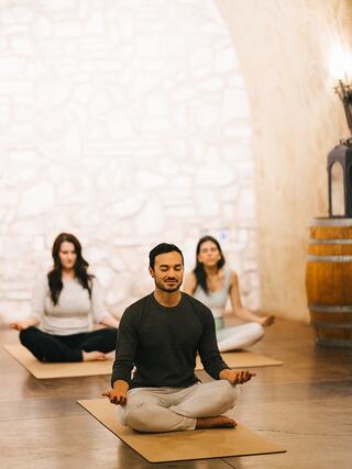 Three individuals practice meditation in a calm, serene environment with stone walls and wooden accents, creating a peaceful atmosphere.