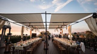 A stylish outdoor dining setup features long tables elegantly set under draped canopies, surrounded by scenic views at sunset.