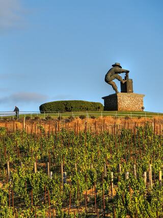 A vineyard stretches out below a large statue of a worker inspecting grapes, set against a clear blue sky.