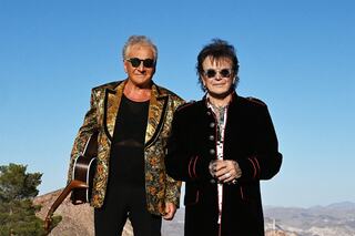 Two musicians stand outdoors against a clear blue sky, one holding a guitar, both in stylish, contrasting outfits. Desert landscape in the background.