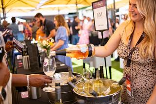 A woman pours wine into a glass at a vibrant outdoor event, surrounded by guests and bottles displayed on tables under sunny umbrellas.