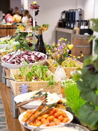A vibrant buffet featuring fresh salads, tomatoes, and colorful flowers, set amidst a warm atmosphere with guests mingling nearby.
