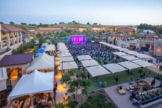 A vibrant outdoor concert scene features a large crowd enjoying live music under tents and twilight skies, surrounded by shops and greenery.