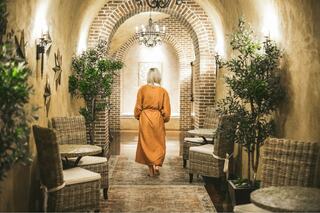A woman in an orange robe walks down a stylish corridor adorned with brick arches, plants, and cozy seating, radiating a warm ambiance.