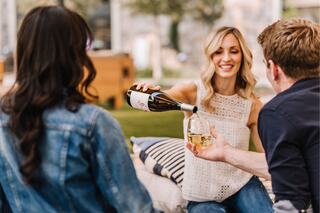 A woman pours wine for a man while sitting outdoors with a friend, enjoying a relaxed social gathering. Laughter and conversation abound.