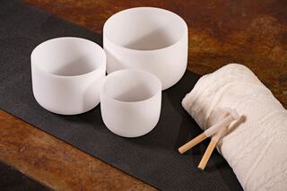 Three white crystal singing bowls of varying sizes sit on a black mat, accompanied by a soft towel and two mallets.
