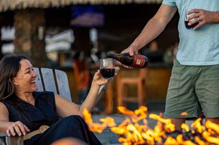 A joyous woman laughs while enjoying a drink, as a man pours another glass by a warm fire, creating a cozy, festive atmosphere.